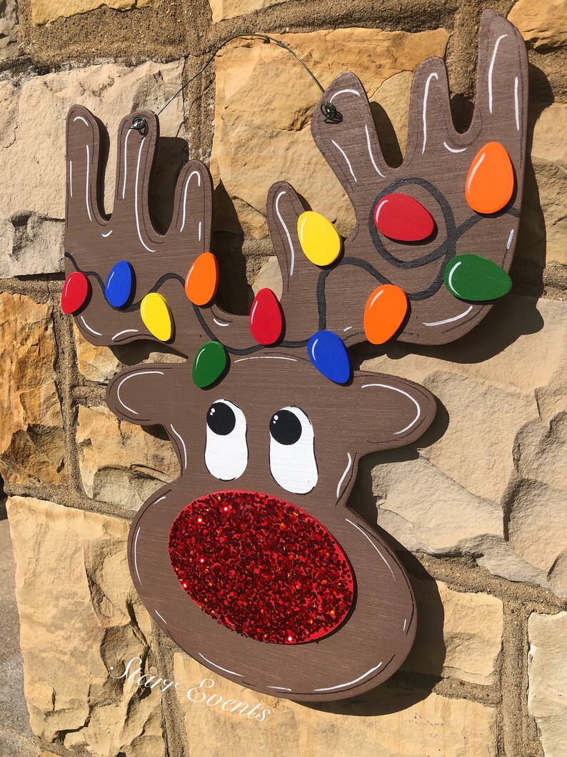 May include: Wooden reindeer Christmas decoration with a brown body, white accents, and a red glitter nose. The antlers are adorned with colorful lights. The decoration is hanging on a stone wall.