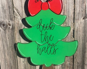 May include: A wooden Christmas tree-shaped sign with a red bow at the top. The sign is painted green with the words "deck the halls" in black script. The tree has a brown trunk and is hanging on a wooden fence.