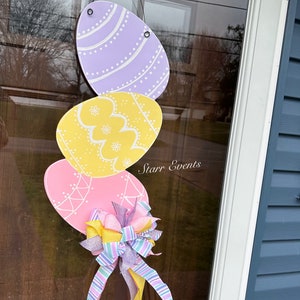 May include: Easter egg door decoration featuring three pastel-colored eggs in lavender, yellow, and pink, adorned with white decorative patterns. A multi-colored bow with ribbons adds a festive touch. The text "Starr Events" is visible.