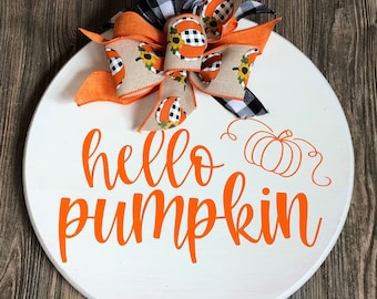 May include: A round, white wooden sign with the words "hello pumpkin" in orange script. A decorative bow with orange, black and white plaid, and sunflower accents sits at the top. The sign is on a wooden background.