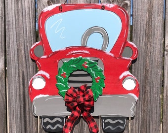 May include: A red wooden truck-shaped door hanger with a light blue windshield and a gray tire. The truck features a green wreath with red berries and a red and black plaid bow. The design is painted on a wooden surface.