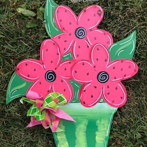 May include: A wooden flower arrangement featuring three pink flowers with black centers and green leaves, set in a green pot. The flowers have a watermelon-like pattern. A pink and green plaid bow adds a decorative touch. The background is grass.