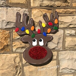 May include: A wooden reindeer decoration with brown antlers adorned with colorful Christmas lights. The reindeer has a red glitter nose and white eyes. The text "Starr Events" is written in cursive below the reindeer.