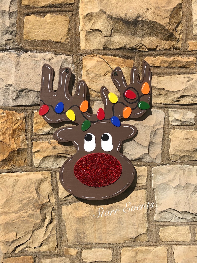 May include: A wooden reindeer decoration with brown antlers adorned with colorful Christmas lights. The reindeer has a red glitter nose and white eyes. The text "Starr Events" is written in cursive below the reindeer.