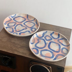 Peut inclure: Deux assiettes en céramique blanche avec un motif abstrait bleu et rose. Les assiettes sont sur une surface en bois.