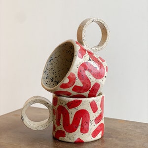 May include: Two ceramic mugs stacked on top of each other. The mugs are off-white with red abstract designs and black speckles. Each mug has a circular handle.