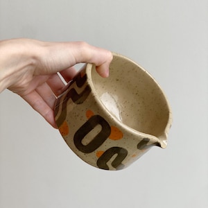 Op de afbeelding: Een gespikkelde beige keramische kom met een tuit, vastgehouden door een hand. De kom is versierd met bruine en oranje abstracte vormen. De binnenkant van de kom is lichtbeige.