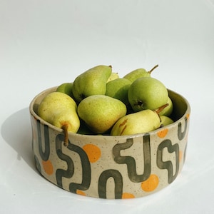 Op de afbeelding: Een keramische kom gevuld met groene peren. De kom heeft een beige achtergrond met een herhalend patroon van donkergroene gebogen lijnen en oranje cirkels. De peren zijn in verschillende tinten groen en geel.