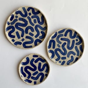 Puede incluir: Tres platos de cerámica de diferentes tamaños. Presentan una base moteada de color blanquecino con un patrón repetitivo de formas azules abstractas. Los platos están dispuestos sobre una superficie blanca.