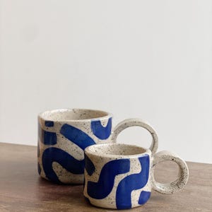 May include: Two ceramic mugs with a white base and blue abstract pattern. The mugs have a speckled glaze and a rounded handle.