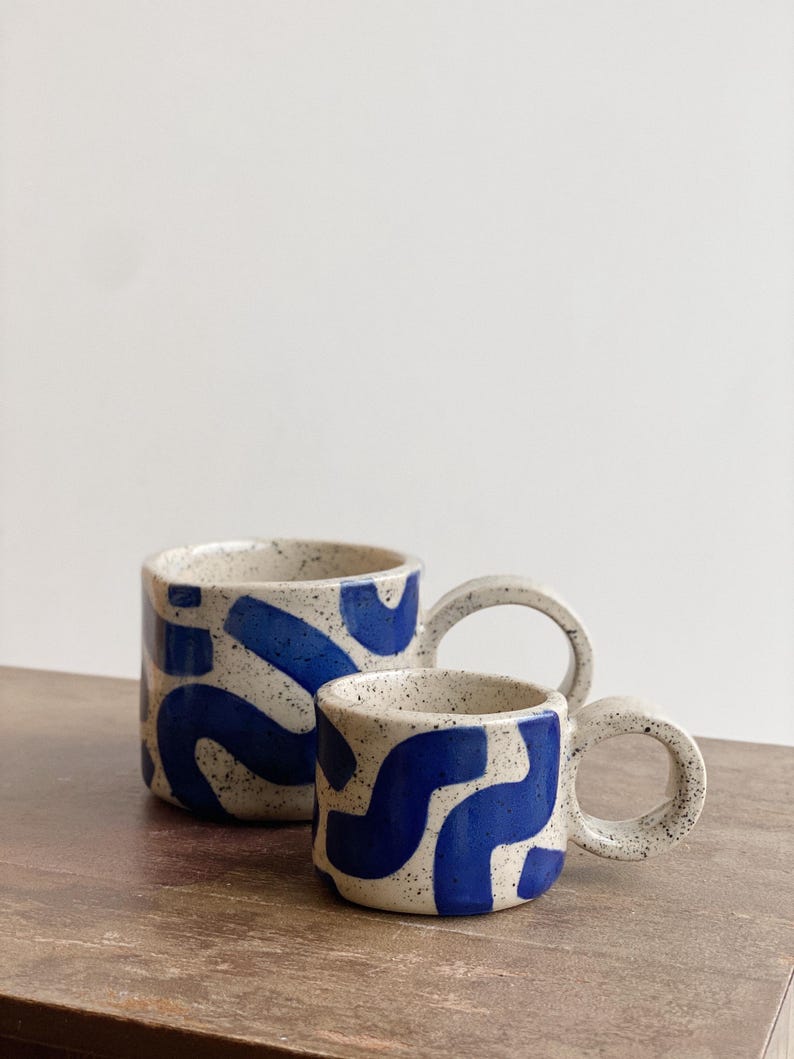 May include: Two ceramic mugs with a white base and blue abstract pattern. The mugs have a speckled glaze and a rounded handle.