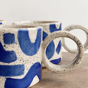 May include: Three ceramic mugs with blue and white abstract designs. The mugs have speckled white glaze and a unique, rounded handle.