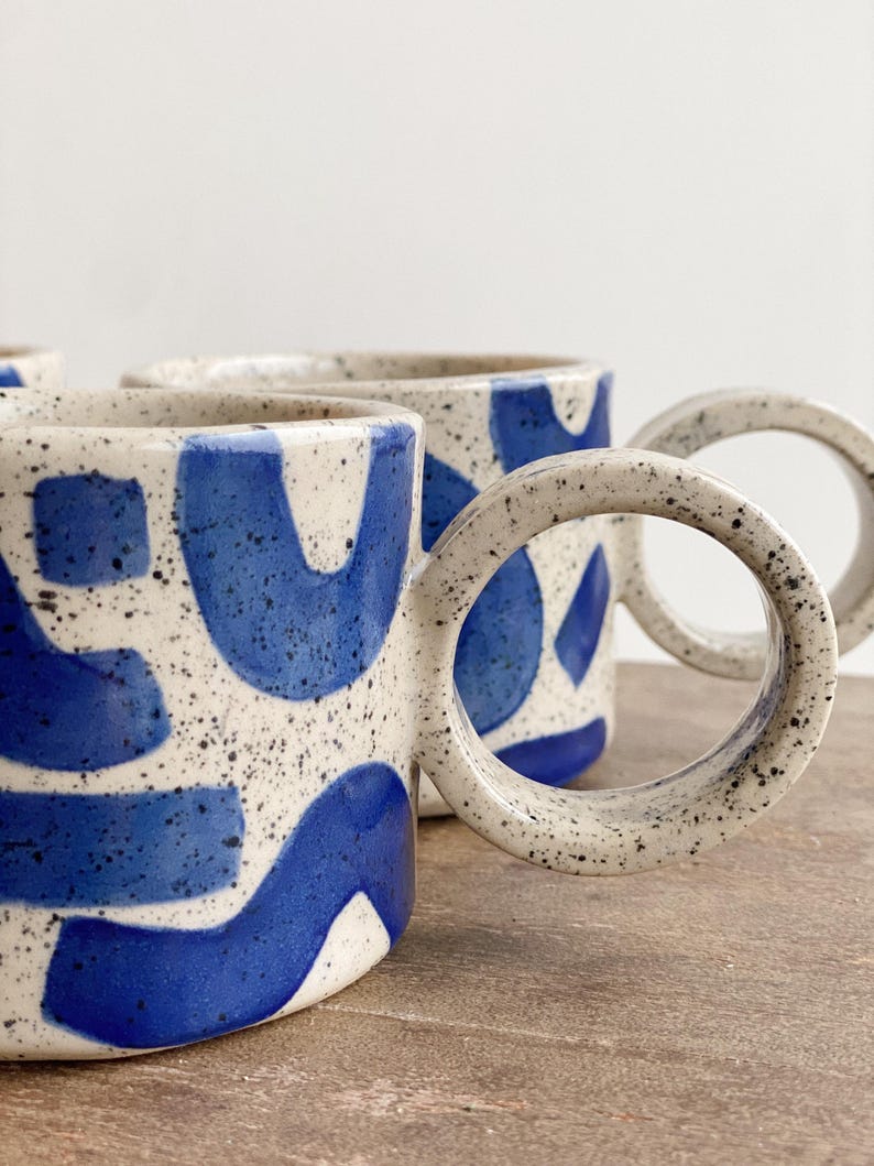 May include: Three ceramic mugs with blue and white abstract designs. The mugs have speckled white glaze and a unique, rounded handle.