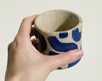 May include: A speckled ceramic mug with a rounded shape, held in a hand. The mug features a cream-colored base with dark speckles and bold blue abstract shapes. The interior is a light cream color.