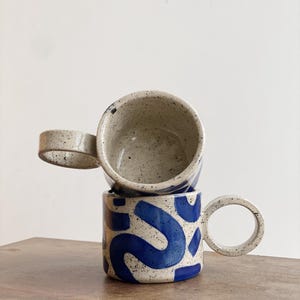 May include: Two ceramic mugs with speckled glaze. The top mug is white with a blue interior. The bottom mug is white with a blue abstract pattern and a large, round handle.