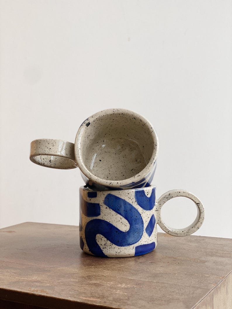 May include: Two ceramic mugs with speckled glaze. The top mug is white with a blue interior. The bottom mug is white with a blue abstract pattern and a large, round handle.