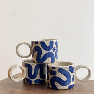 May include: Three ceramic mugs with blue and white abstract designs and large, round handles.