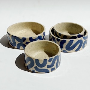 Pode incluir: Três tigelas redondas de cerâmica com uma base creme salpicada e um padrão abstrato de rabiscos azul-escuro pintado à mão no exterior. As tigelas variam em tamanho e apresentam uma forma cilíndrica lisa com uma base plana.