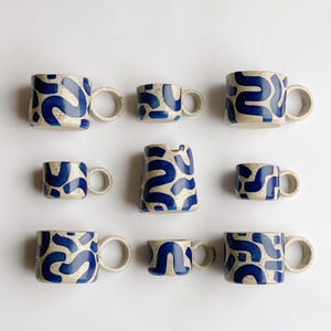 May include: Set of nine ceramic mugs in various sizes, all featuring a white background with a blue abstract pattern. The mugs have a rounded shape and a thick, rounded handle.