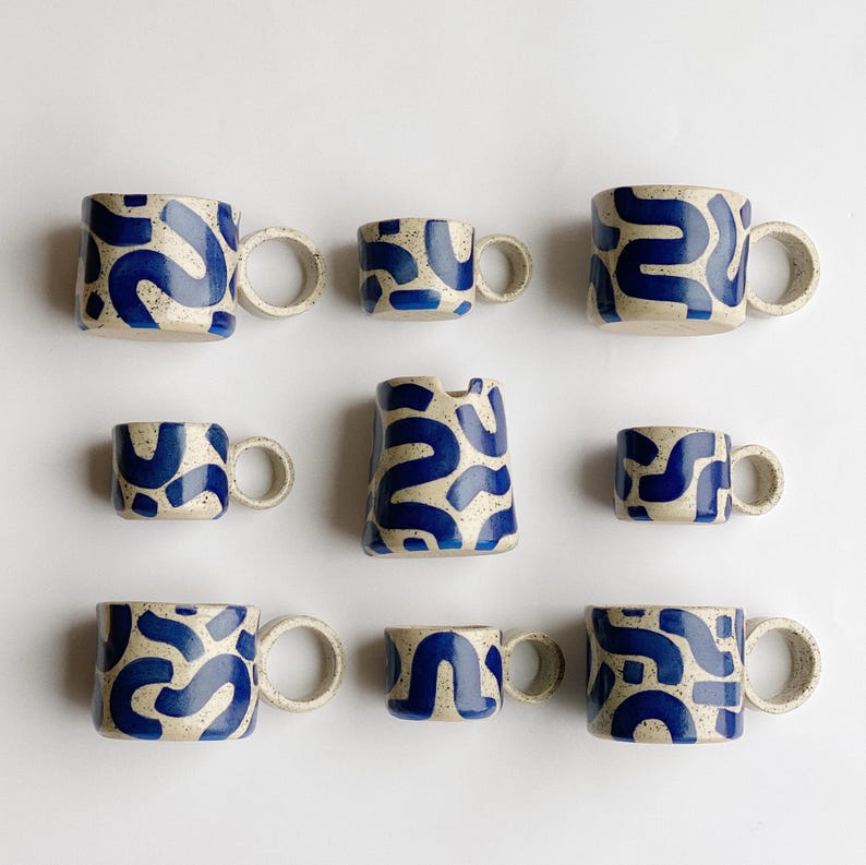 May include: Set of nine ceramic mugs in various sizes, all featuring a white background with a blue abstract pattern. The mugs have a rounded shape and a thick, rounded handle.