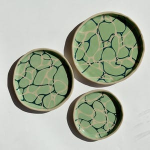 May include: Three ceramic plates with a marbled design. The plates are in varying sizes, with a light green background and dark green and off-white veining. The rims are a light beige colour.