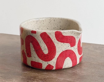 May include: A small, speckled ceramic bowl with a red abstract design. The bowl has a rounded shape with a small spout. The exterior features a cream-colored base with black speckles and a bold red pattern.