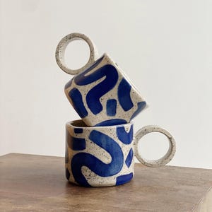 May include: Two ceramic mugs with blue and white abstract patterns. The mugs have unique, circular handles.