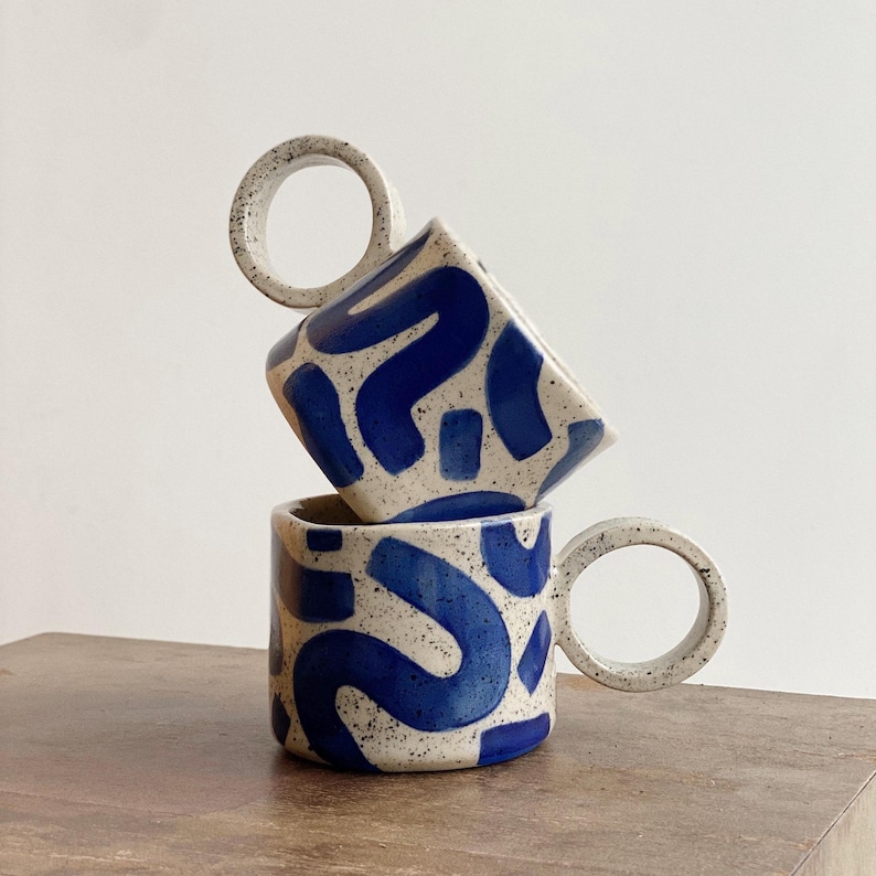 May include: Two ceramic mugs with blue and white abstract patterns. The mugs have unique, circular handles.