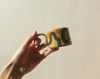 Könnte beinhalten: Eine beige Keramiktasse mit einem einzigartigen abstrakten Design aus schwarzen und orangefarbenen Linien und Kreisen. Die Tasse hat einen abgerundeten Henkel.