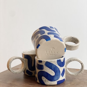 May include: Three ceramic mugs with blue and white abstract patterns. The mugs have large, rounded handles. The bottom mug has the text "Tilia Studio" on the bottom.