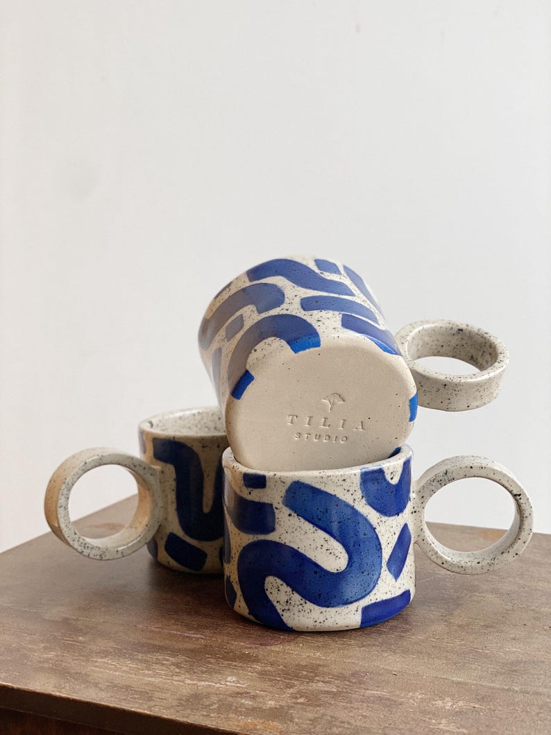 May include: Three ceramic mugs with blue and white abstract patterns. The mugs have large, rounded handles. The bottom mug has the text "Tilia Studio" on the bottom.