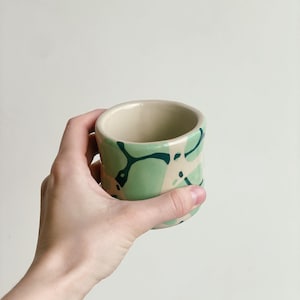 May include: A green and white ceramic mug with a swirling abstract design. The mug is held in a hand.