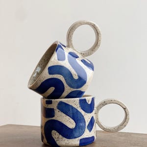 May include: Two ceramic mugs with blue and white abstract patterns and unique circular handles. The mugs are stacked on top of each other.