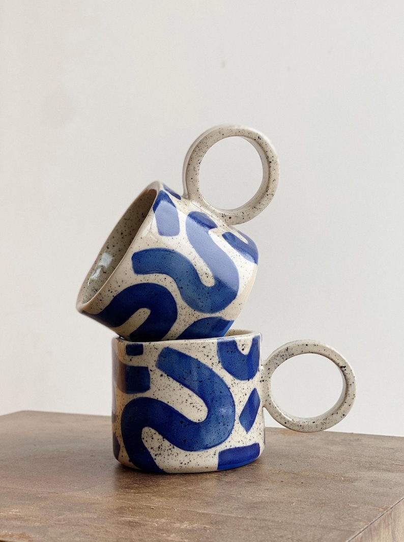 May include: Two ceramic mugs with blue and white abstract patterns and unique circular handles. The mugs are stacked on top of each other.