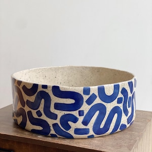 May include: A ceramic bowl with a speckled beige interior and exterior. The exterior features a repeating pattern of blue abstract shapes. The bowl is round and sits on a wooden surface.