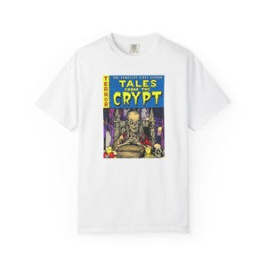 May include: A white short-sleeve t-shirt featuring a graphic print of the Tales from the Crypt comic book cover. The design displays a skeletal figure sitting behind a large book with yellow text reading TERROR TALES FROM THE CRYPT.