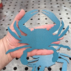 May include: A blue metal crab cutout, held in a hand, with a textured surface. The crab is a popular nautical theme for home decor.