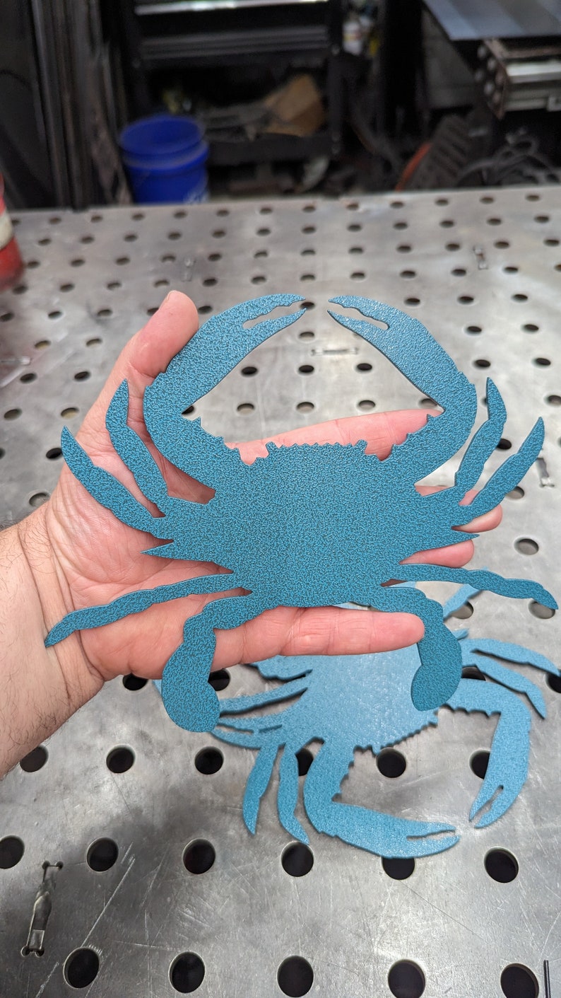May include: A blue metal crab cutout, held in a hand, with a textured surface. The crab is a popular nautical theme for home decor.