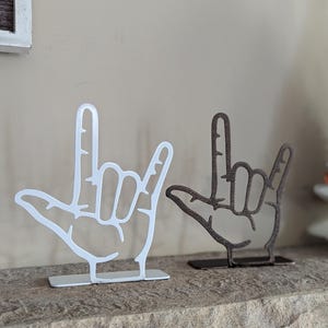 May include: Two metal sculptures of hands forming the American Sign Language symbol for "I love you". One sculpture is white and the other is brown. The sculptures are on a wooden shelf.