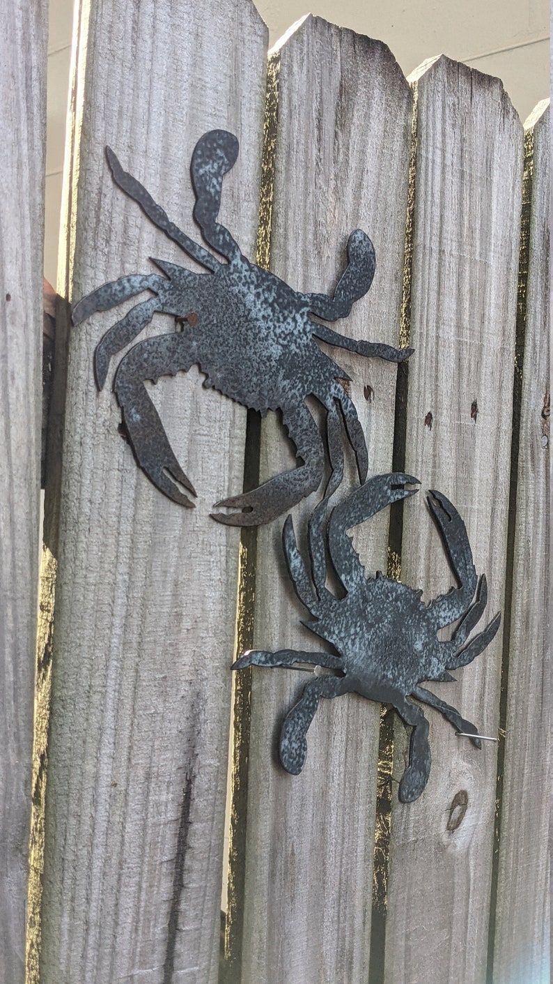 May include: Two metal crab silhouettes attached to a wooden fence. The crabs are painted a dark gray with a weathered finish.