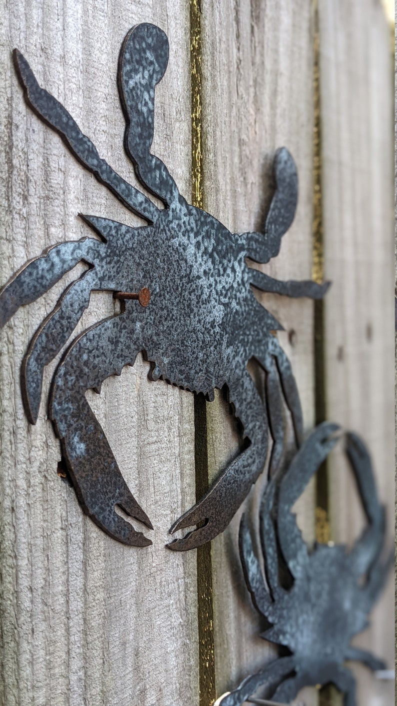 May include: Two metal crab silhouettes, one larger than the other, are attached to a wooden fence. The larger crab is in the foreground and is facing the viewer. The smaller crab is in the background and is facing the right. Both crabs are painted a dark gray color.