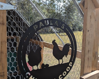 May include: A black metal sign for a chicken coop, with the words "Paula's Chicken Coop" cut out in a circular design. The sign features silhouettes of a hen, a rooster, and eggs. The sign is hanging on a wooden frame with chicken wire.