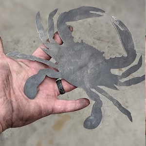 May include: A metal cutout of a crab in a hand. The crab is a silhouette with a detailed design. The crab is gray in color.