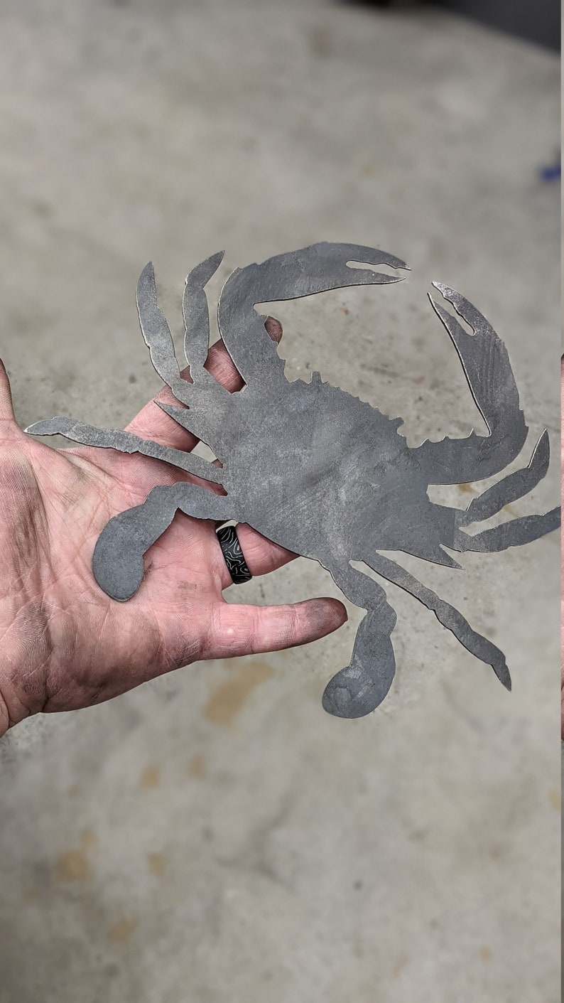 May include: A metal cutout of a crab in a hand. The crab is a silhouette with a detailed design. The crab is gray in color.