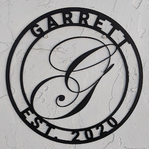 May include: Black metal circular sign with the name "GARRETT" and the year "EST. 2020" around the outside. A large, stylised letter "G" is in the centre.
