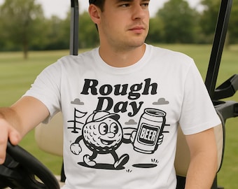 Pode incluir: Camiseta branca com um personagem de desenho animado de uma bola de golfe com um chapéu e uma lata de cerveja. O texto "Rough Day" está acima do gráfico. A bola de golfe está andando para a direita.
