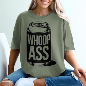 Puede incluir: Camiseta verde oliva con un gráfico negro de una lata de refresco con las palabras "WHOOP ASS" en letras blancas. La camiseta es de estilo casual y unisex, adecuada para el uso diario.