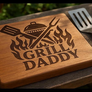 Puede incluir: Una tabla de cortar de madera con las palabras "GRILL DADDY" grabadas, junto con un diseño de parrilla y llamas. Una espátula de metal descansa junto a la tabla. La tabla es de madera.