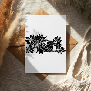 Op de afbeelding: Een witte wenskaart met een zwarte illustratie van een tak met bladeren, een kokosnoot en een pot honing. De kaart ligt op een bruine envelop, met een beige achtergrond en een witte draagtas.