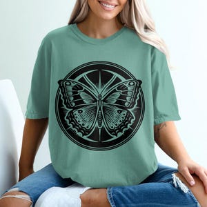 May include: A teal blue t-shirt with a black and white graphic of a butterfly inside a circle.